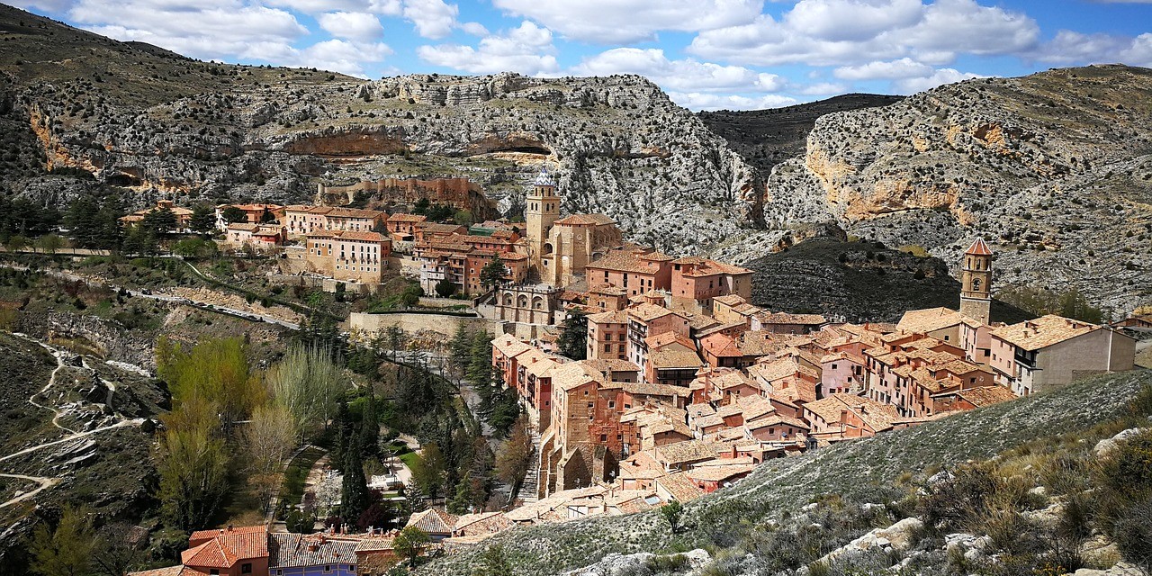 Aerial view of Albarracín, Spain, showing terracotta-roofed mediaeval buildings nestled in a rocky valley surrounded by mountains.