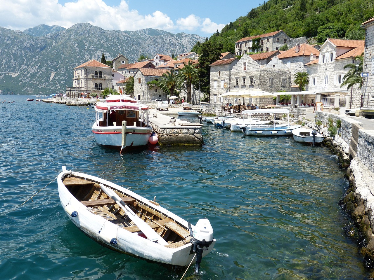Waterfront view of Kotor, Montenegro, with stone houses, moored boats, and mountains in the background under a clear sky.