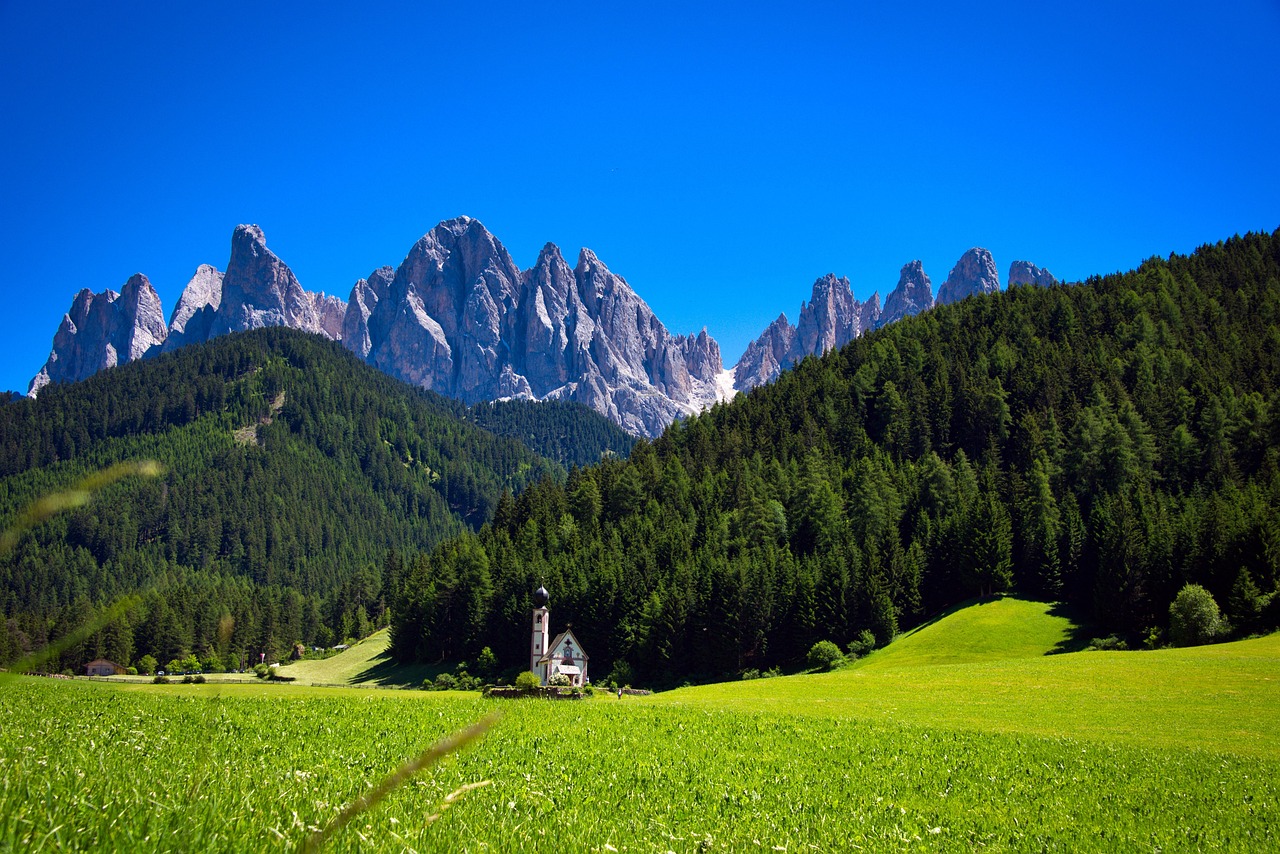  A bright green meadow in the foreground with a small white church with a tall steeple in the middle ground. Behind the church are rolling hills covered in a dense pine forest, and behind the forest, a jagged range of rocky, grey mountains stand against a clear blue sky.