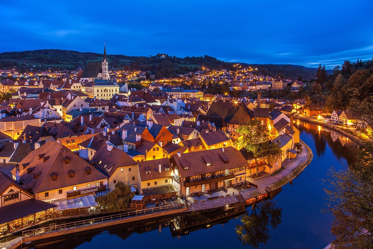 An aerial, dusk photo of a charming old town on the banks of a river. The town's buildings are lit up with warm yellow lights. In the distance, a large church steeple with a clock face rises above the rest of the buildings, and beyond that, rolling hills are visible against a dark blue sky.
