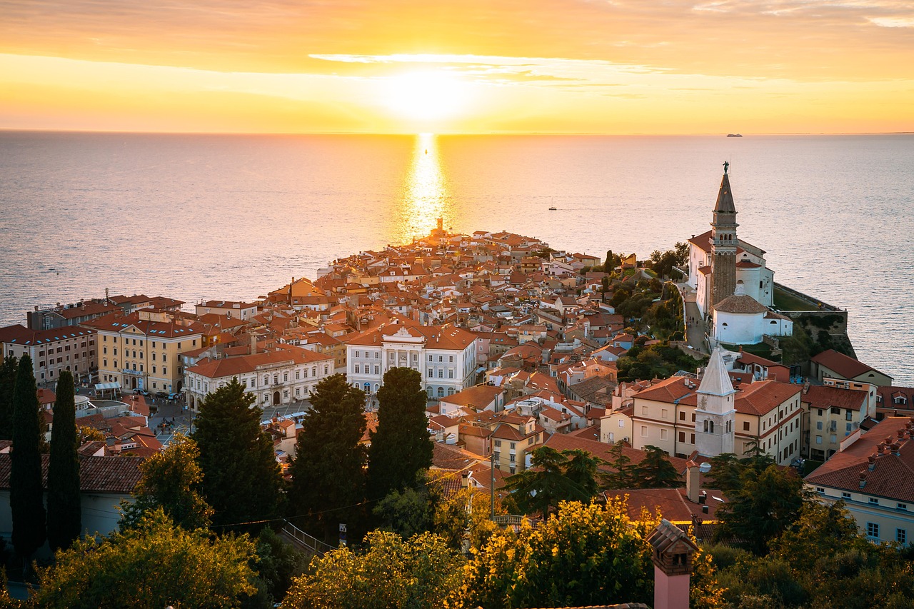  An aerial view of a coastal town during sunset. The town, with its red-roofed buildings, is situated on a peninsula jutting out into the sea. The sun, a bright yellow light, is setting on the horizon, creating a sunbeam on the water. A tall, white church steeple with a clock face rises from the town, surrounded by other buildings.