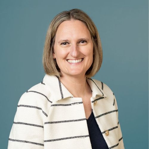 Smiling woman with shoulder-length blonde hair wearing a white jacket with black stripes and a black top.