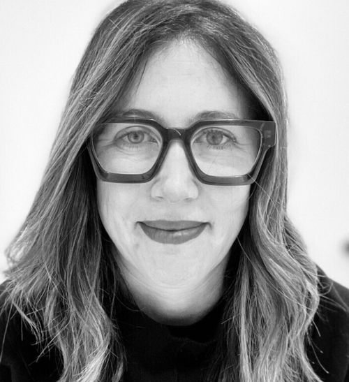 Close-up of a woman with long hair and large glasses smiling slightly, black and white photo.