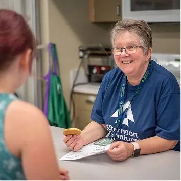 Smiling woman with glasses offers a cookie and paper to another woman at a counter.