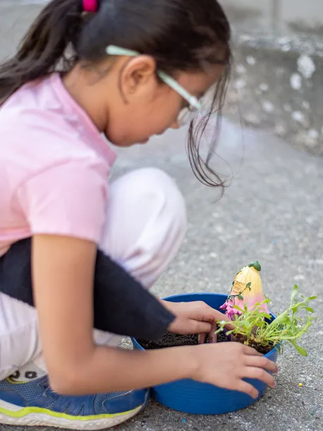 Child in glasses planting a small green plant in a blue pot outdoors on concrete.