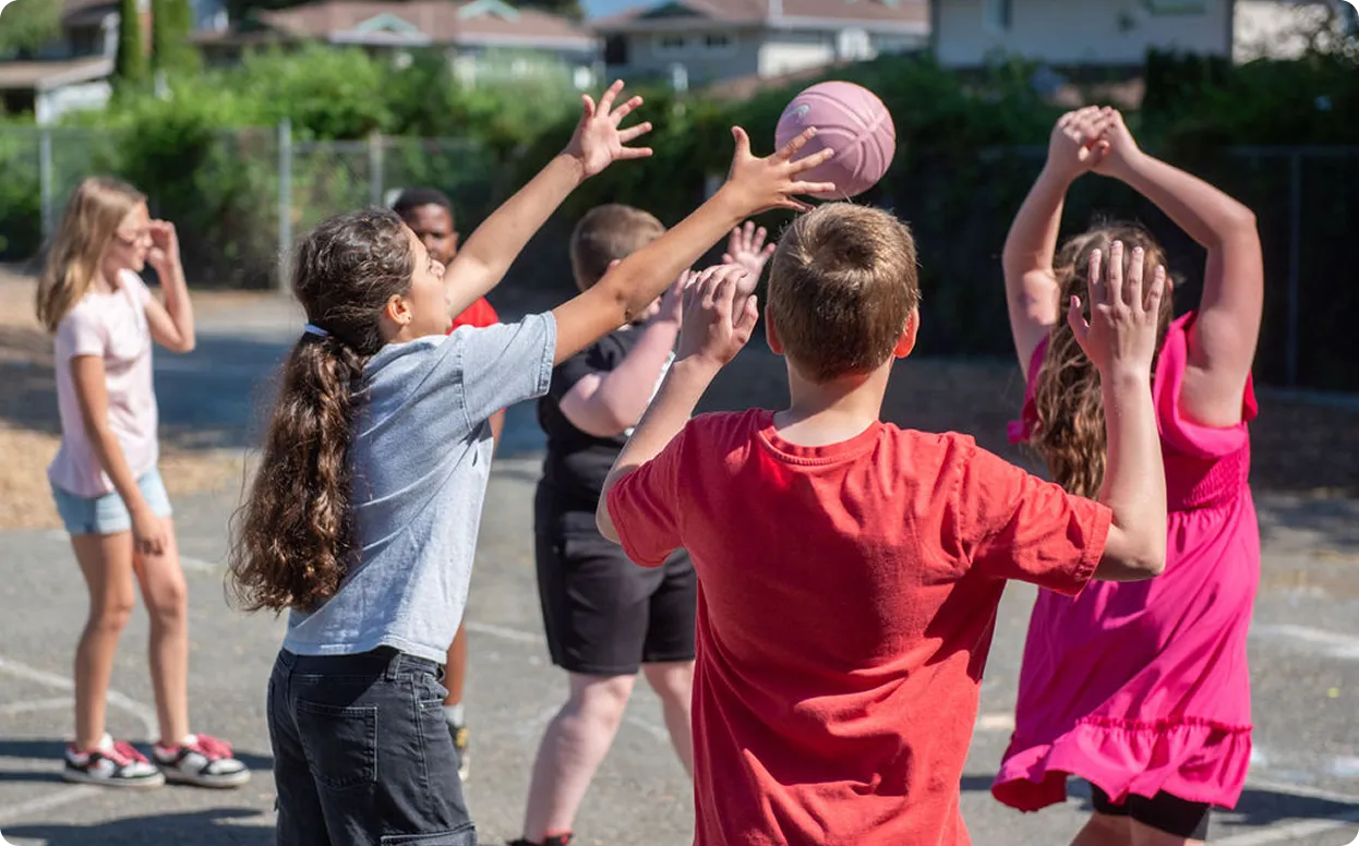 Children playing basketball outdoors, one girl reaching to catch a pink ball midair on a sunny day.