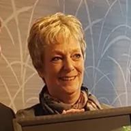 Smiling older woman with short blonde hair and scarf against a light patterned background.