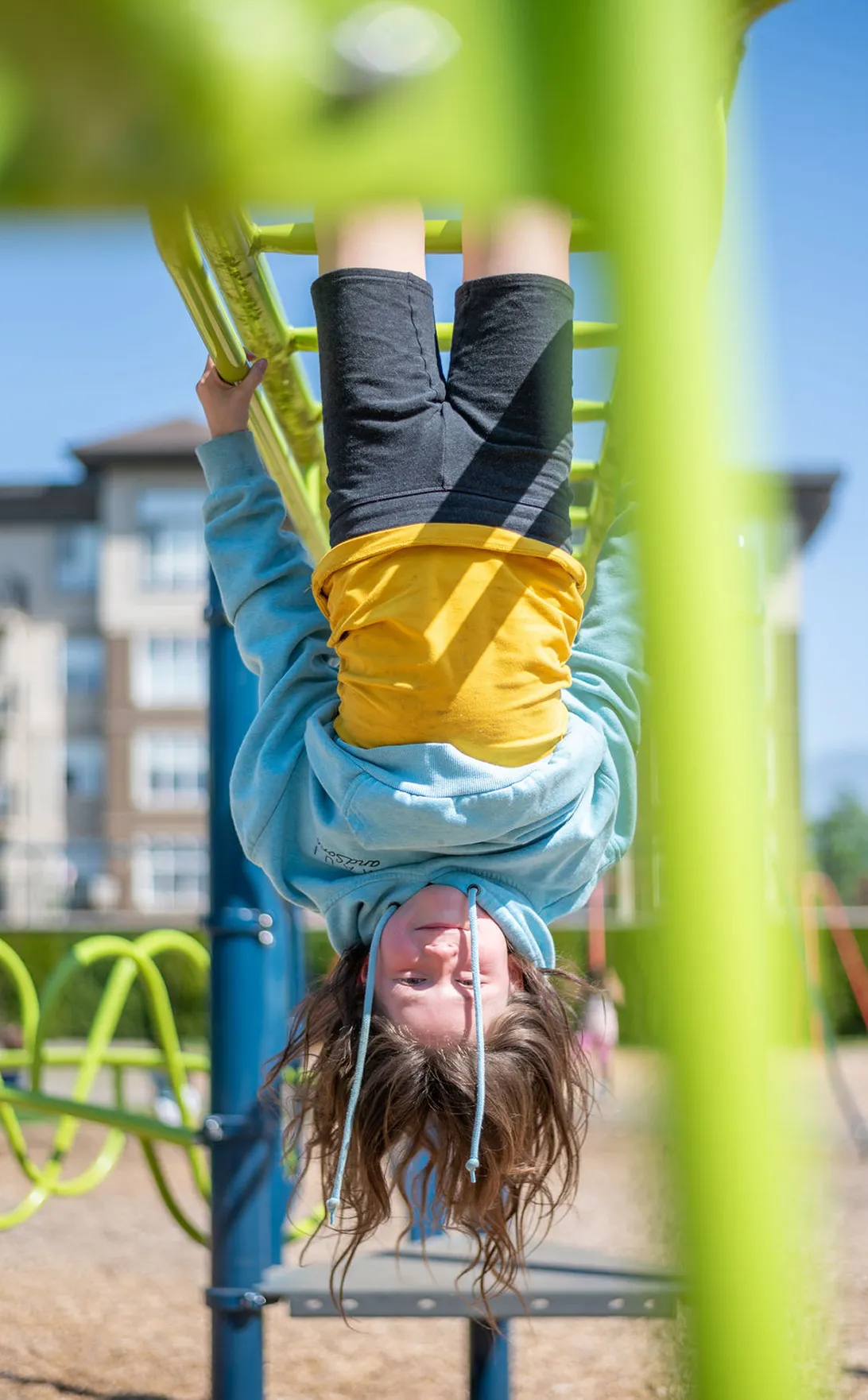 Child hanging upside down from green monkey bars at a playground on a sunny day.