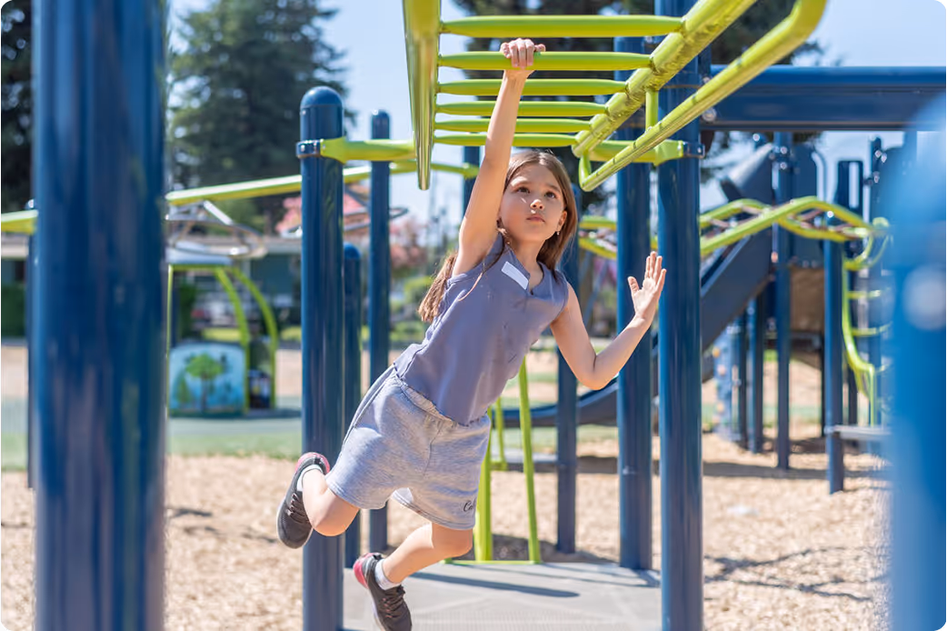 Girl with long hair wearing blue and gray plays on outdoor monkey bars at playground.