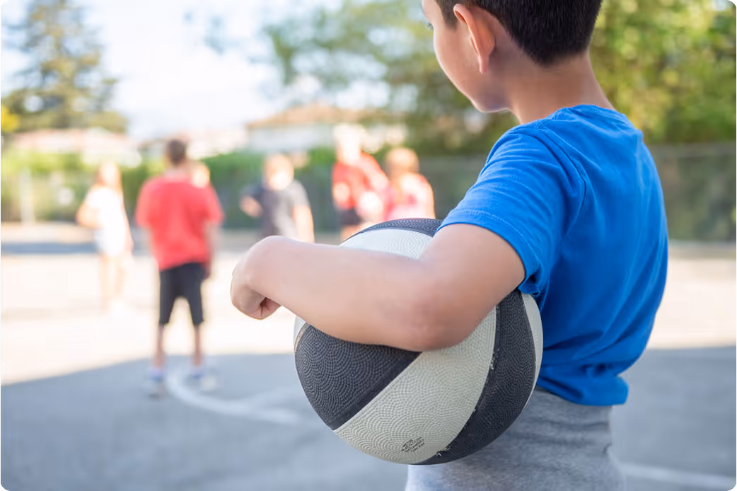 Young boy in blue shirt holding a basketball, with other children playing in the background.