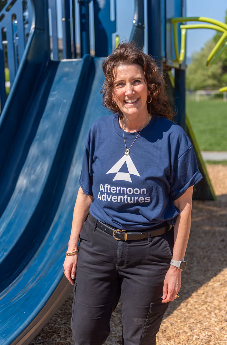 Smiling woman wearing a navy Afternoon Adventures shirt stands in front of a blue playground slide.