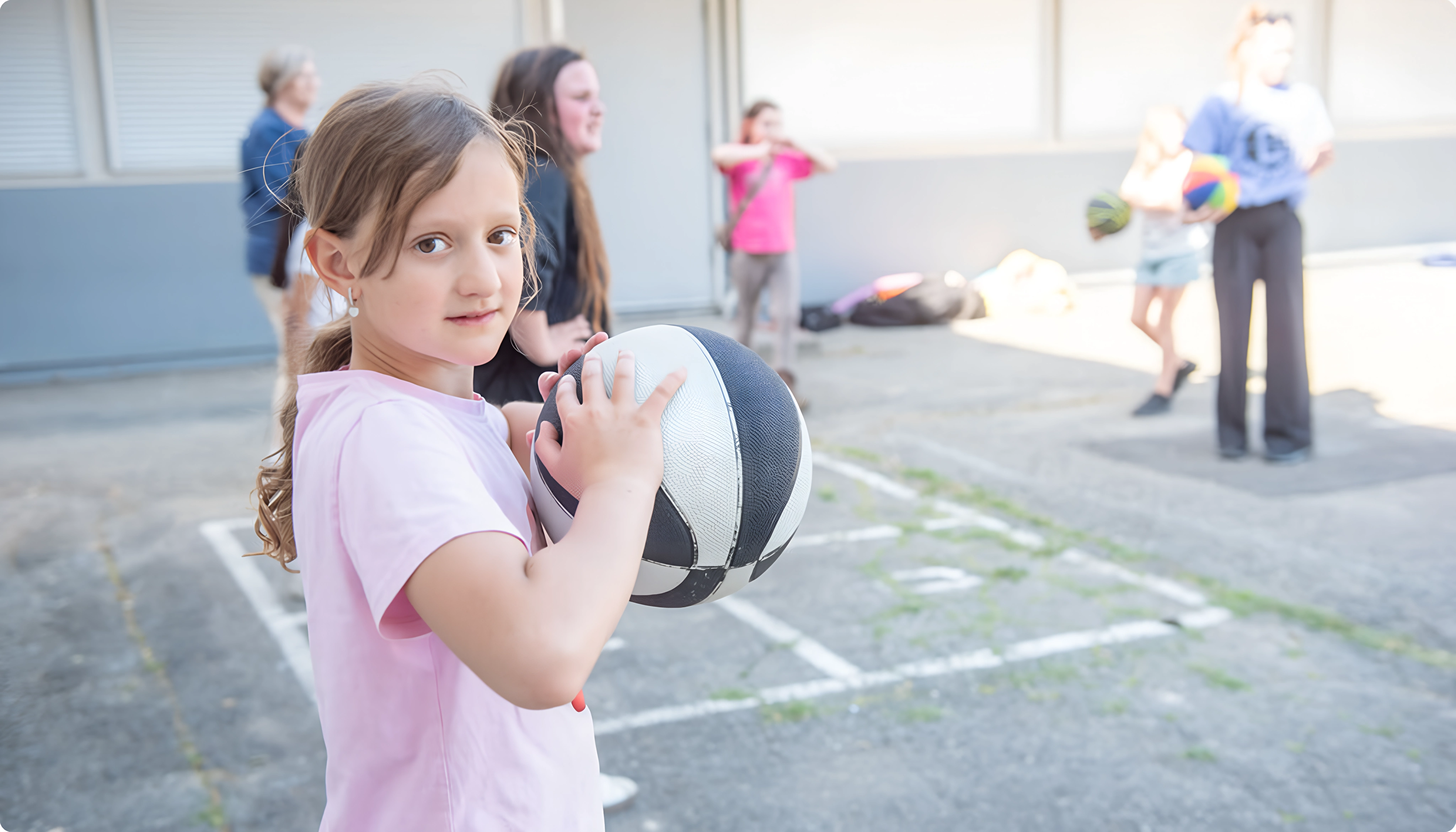 Young girl in pink shirt holding a black and white basketball outdoors with other kids playing.