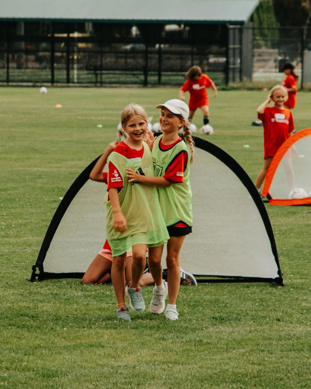 Two young girls in green vests smiling and hugging on a soccer field during practice.