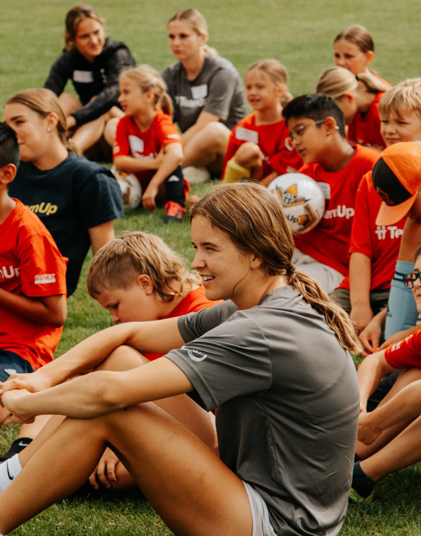 Group of children and coaches sitting on grass at a soccer camp, wearing red and gray shirts.