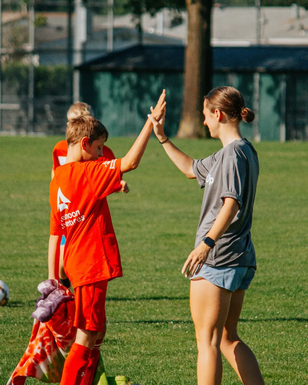 Child soccer player in red high-fiving adult coach in gray shirt on green field.