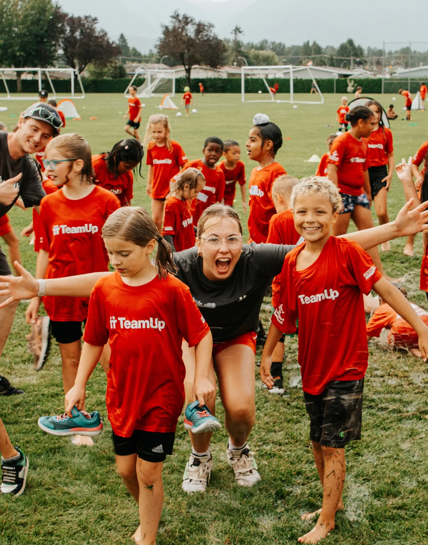 Smiling kids and an adult in red TeamUp shirts on grassy soccer field with goals in background.