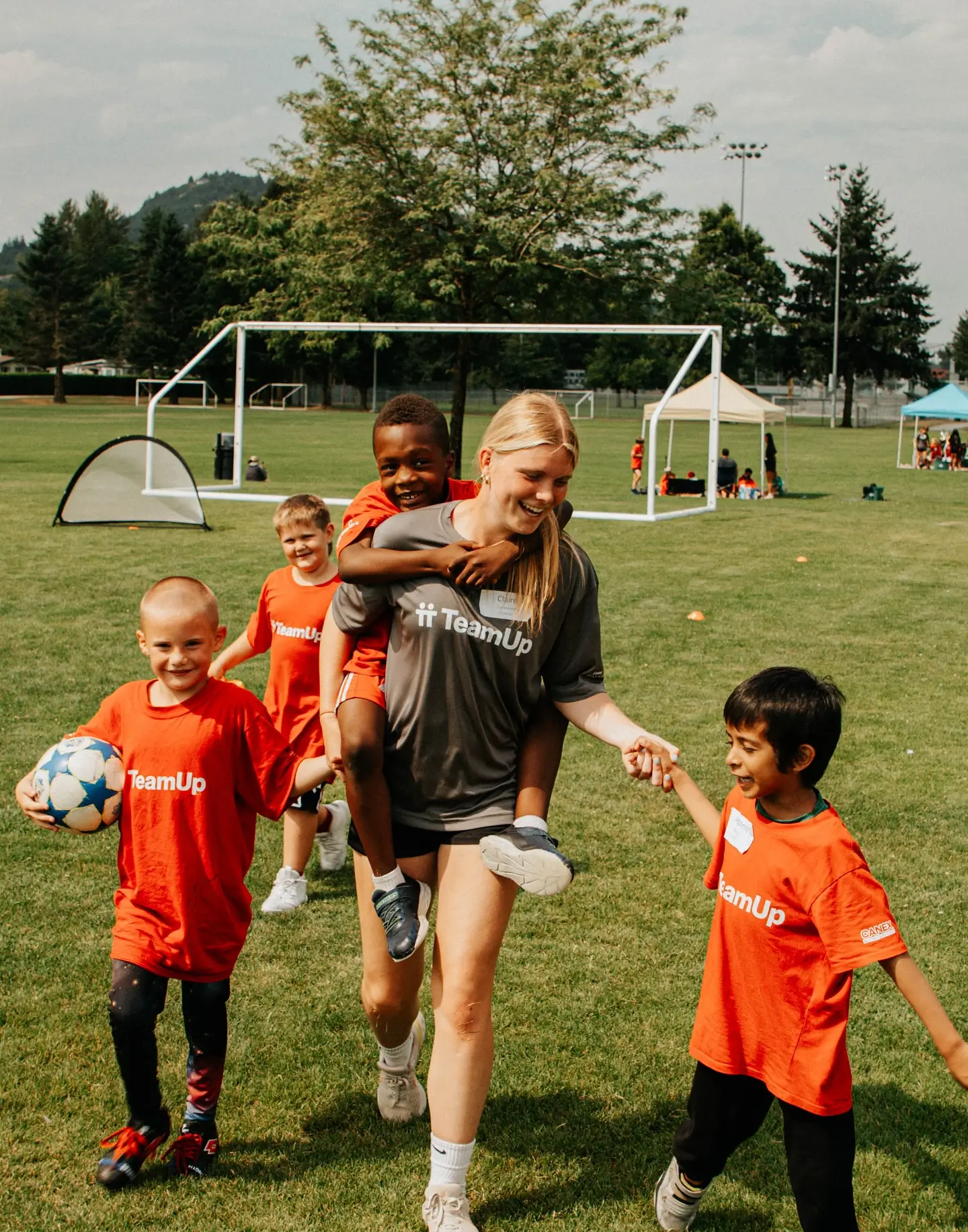 Young woman carrying a boy on her back while holding hands with two other children in orange TeamUp shirts on a soccer field.
