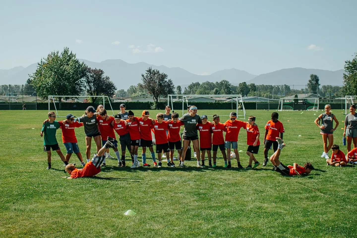 Kids and coaches in TeamUp shirts standing arm in arm on a soccer field with goals behind them.