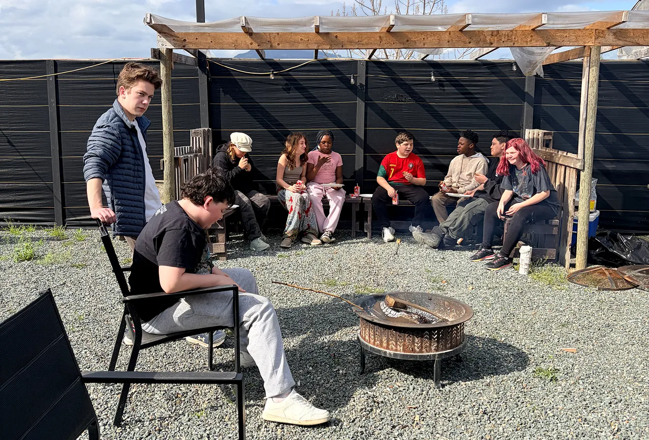 Group of young people sitting and standing around a fire pit in an outdoor gravel area under a wooden pergola.