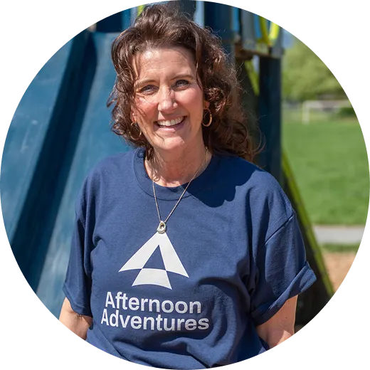 Smiling woman wearing a navy Afternoon Adventures t-shirt outdoors near playground equipment.