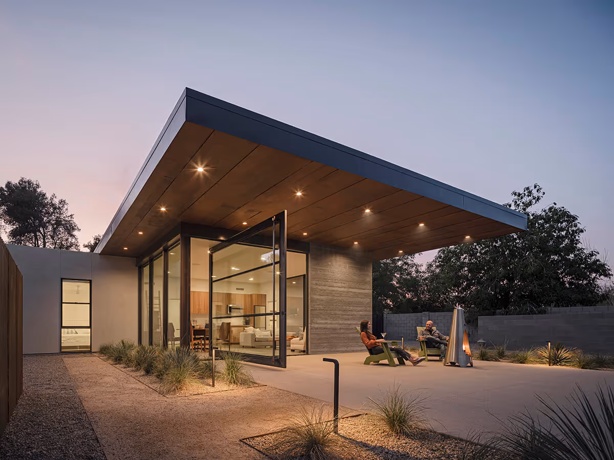 Dusk view of a huge steel and glass pivot door, large cantilevered roof with wood ceiling and boardformed concrete wall and stucco on a casita in Phoenix by The Ranch Mine