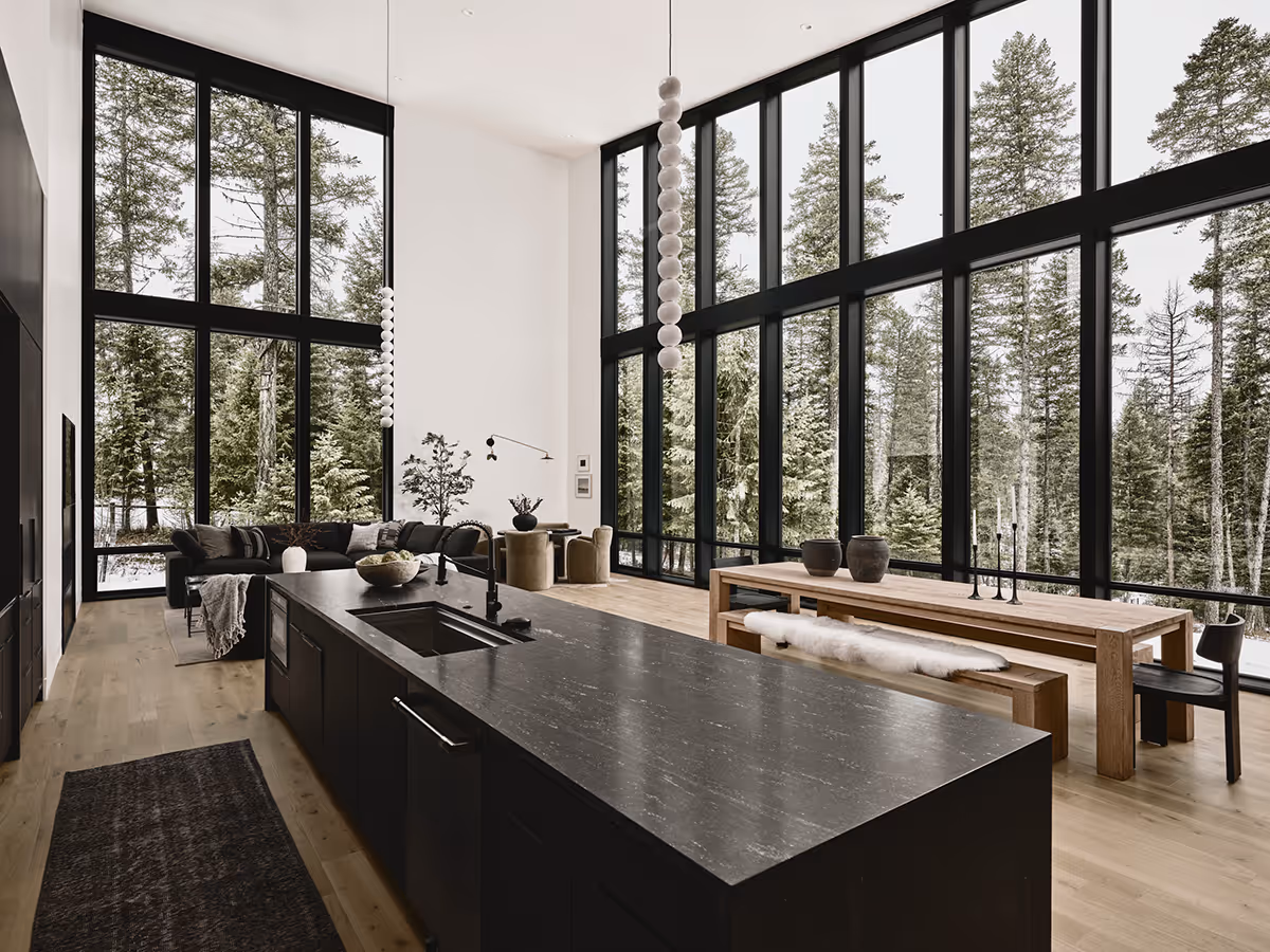 Great room with a black island and lots of windows looking at a pine forest in Whitefish, Montana
