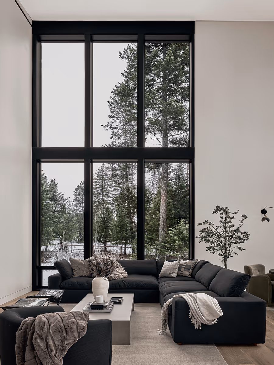 Living room with a gray sofa and lots of windows looking at a pine forest in Whitefish, Montana