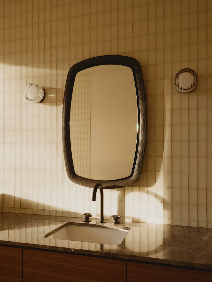 Wood-framed mirror above a stone vanity in a cream tile bath, morning light raking across the tile