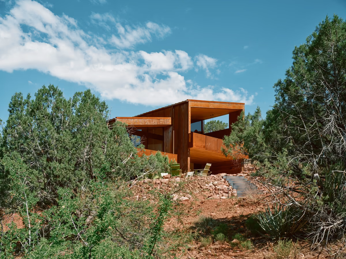 Weathering steel guest house set among juniper on a rocky slope, lounge chairs on the terrace below