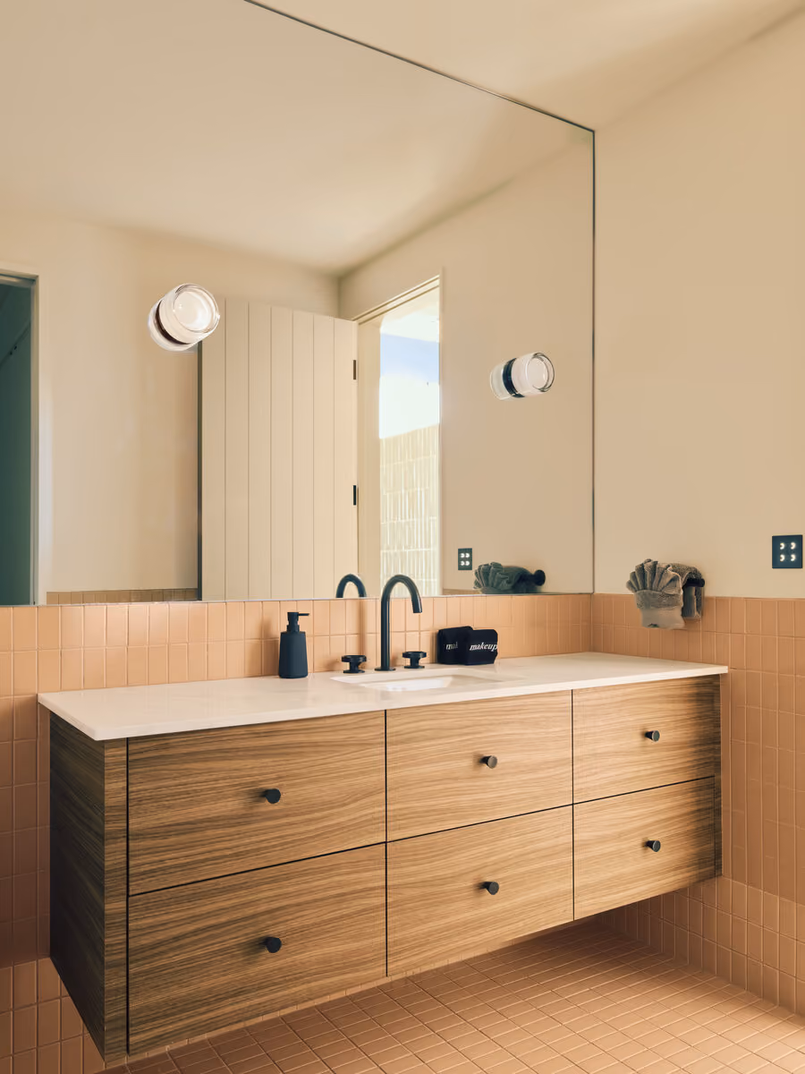Floating walnut vanity above blush pink tile with a full-width mirror and black fixtures