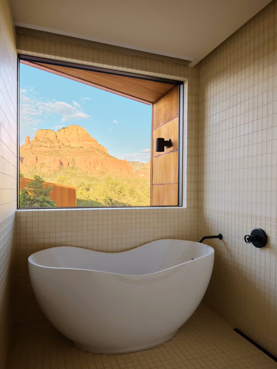 Freestanding soaking tub set below a square window framing a red rock butte in a white tile bath