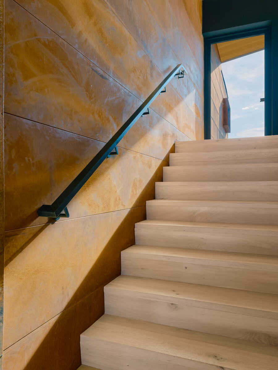 White oak stair rising against a weathering steel wall with a black handrail