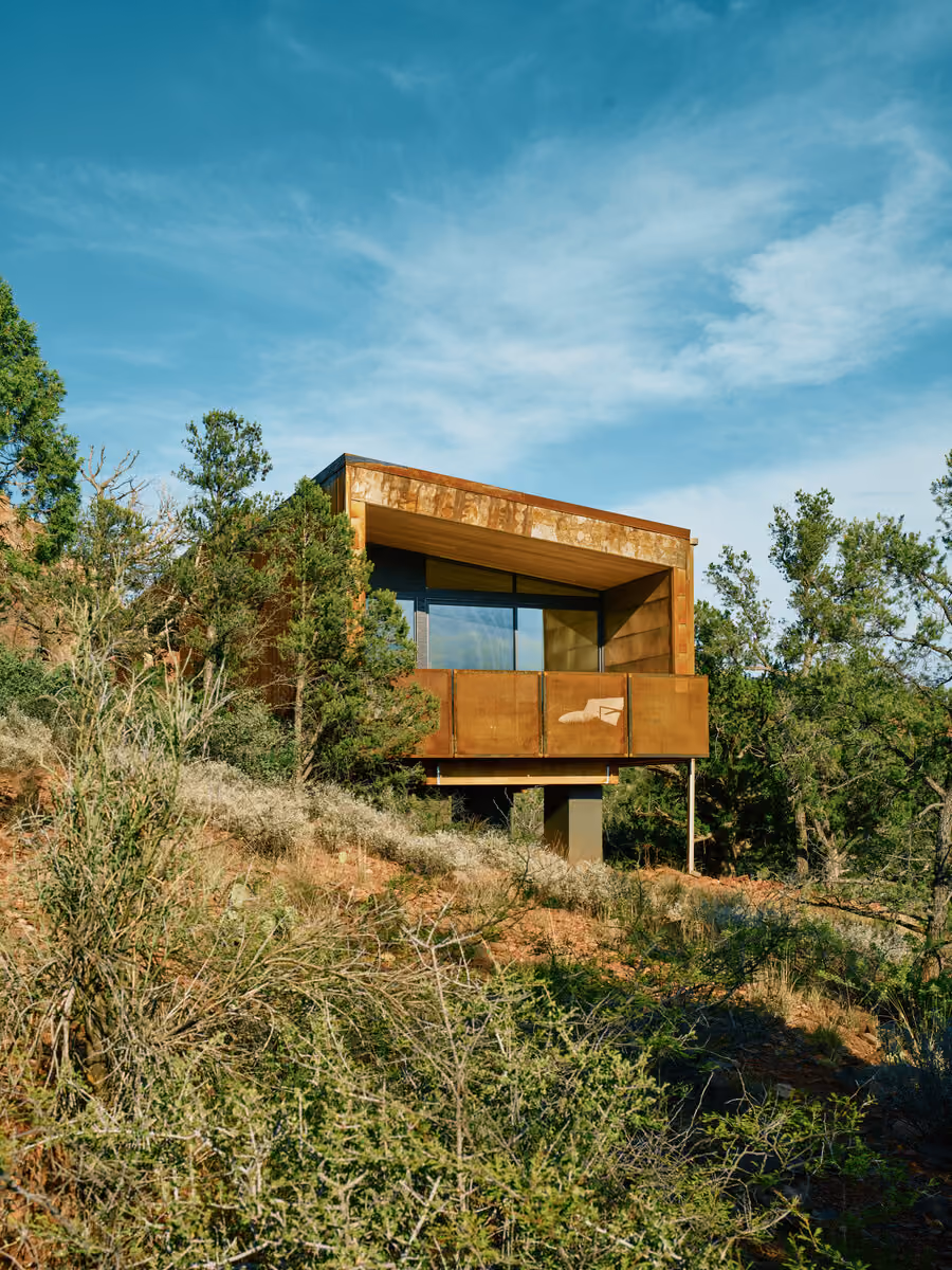 Cabin cantilevered on a concrete pier above a juniper hillside, corten balcony facing the view