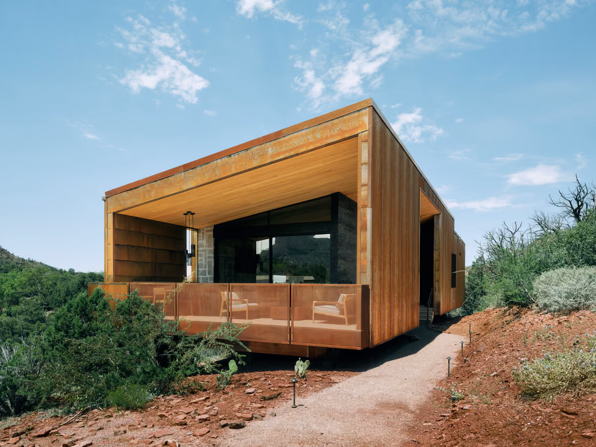 Corten cabin with a deep covered porch and stone-walled interior visible through the glass