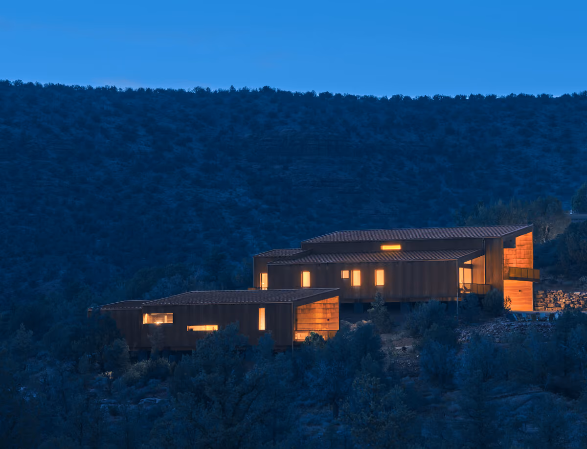 Both homes lit from within at blue hour, stepping down a dark juniper hillside