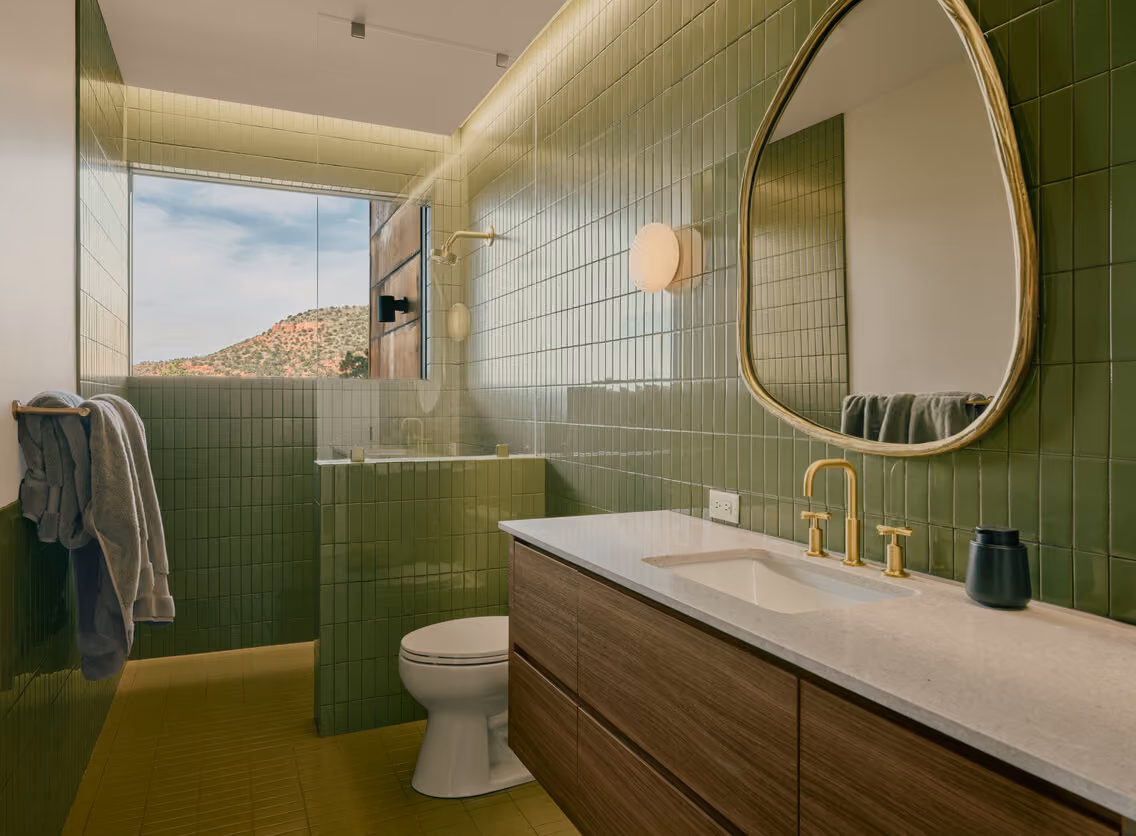Green tile bath with a brass-framed mirror, walnut vanity and a window onto the red rock hillside