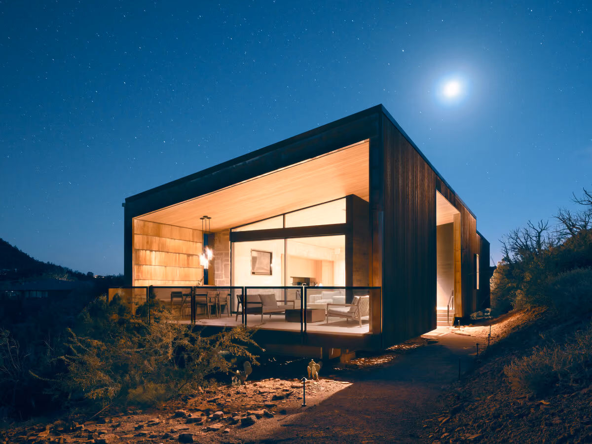 The cabin lit from within under a full moon and stars, glass wall open to the desert