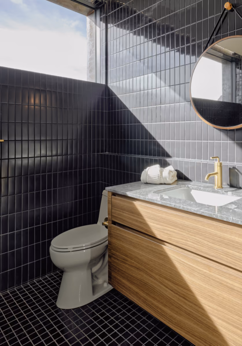 Guest bath in glossy black tile with brass fixtures, a round mirror and a white oak vanity