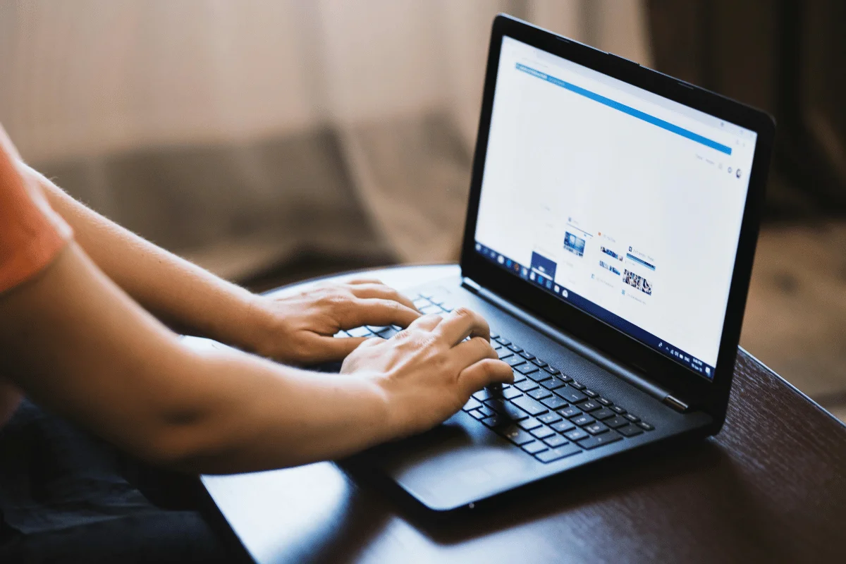 A person types on a black laptop with a webpage open, sunlight streaming through a nearby window. The scene conveys focus and productivity.