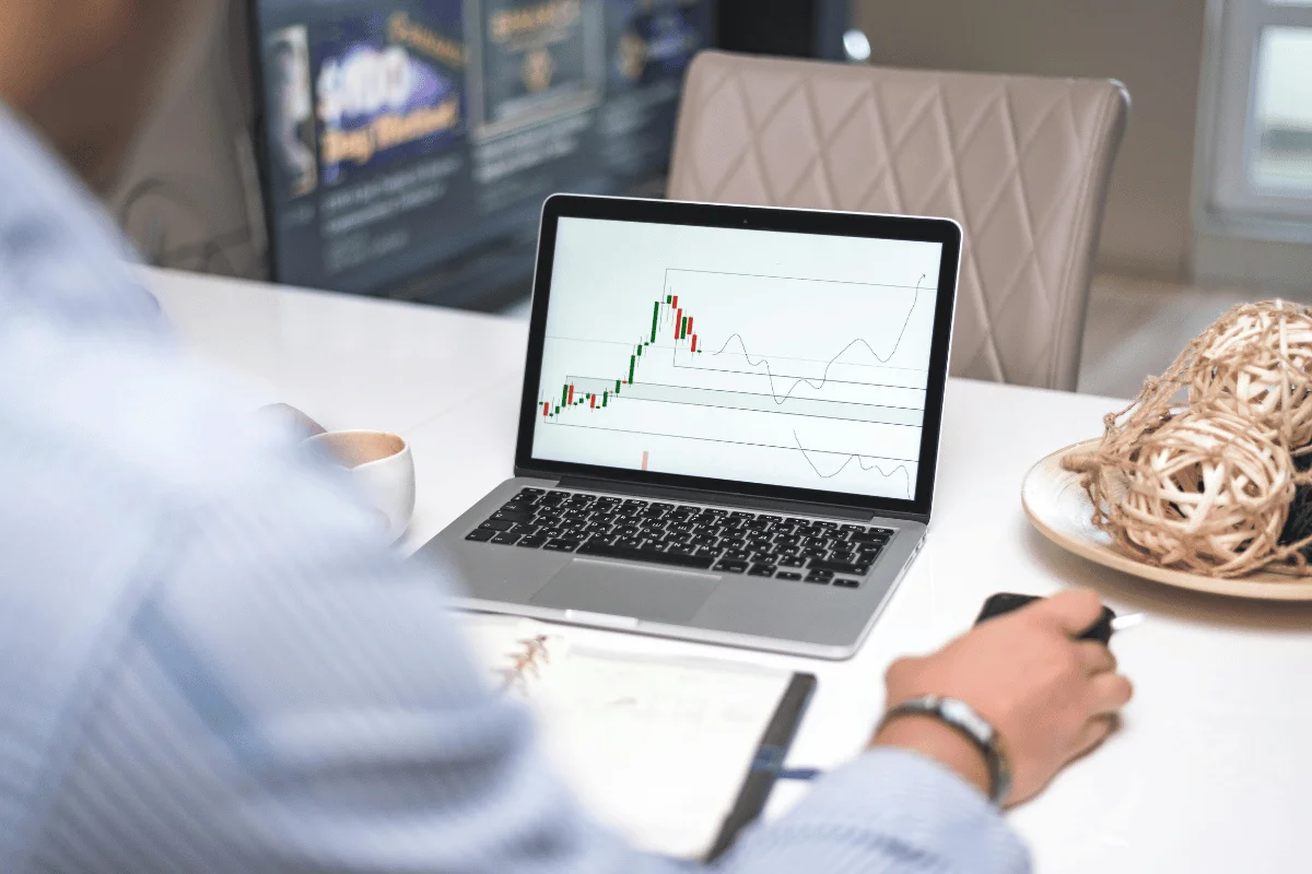 A person analyzes a stock market graph on a laptop screen at a desk. The scene is professional and focused, with a coffee cup and decorative item nearby.