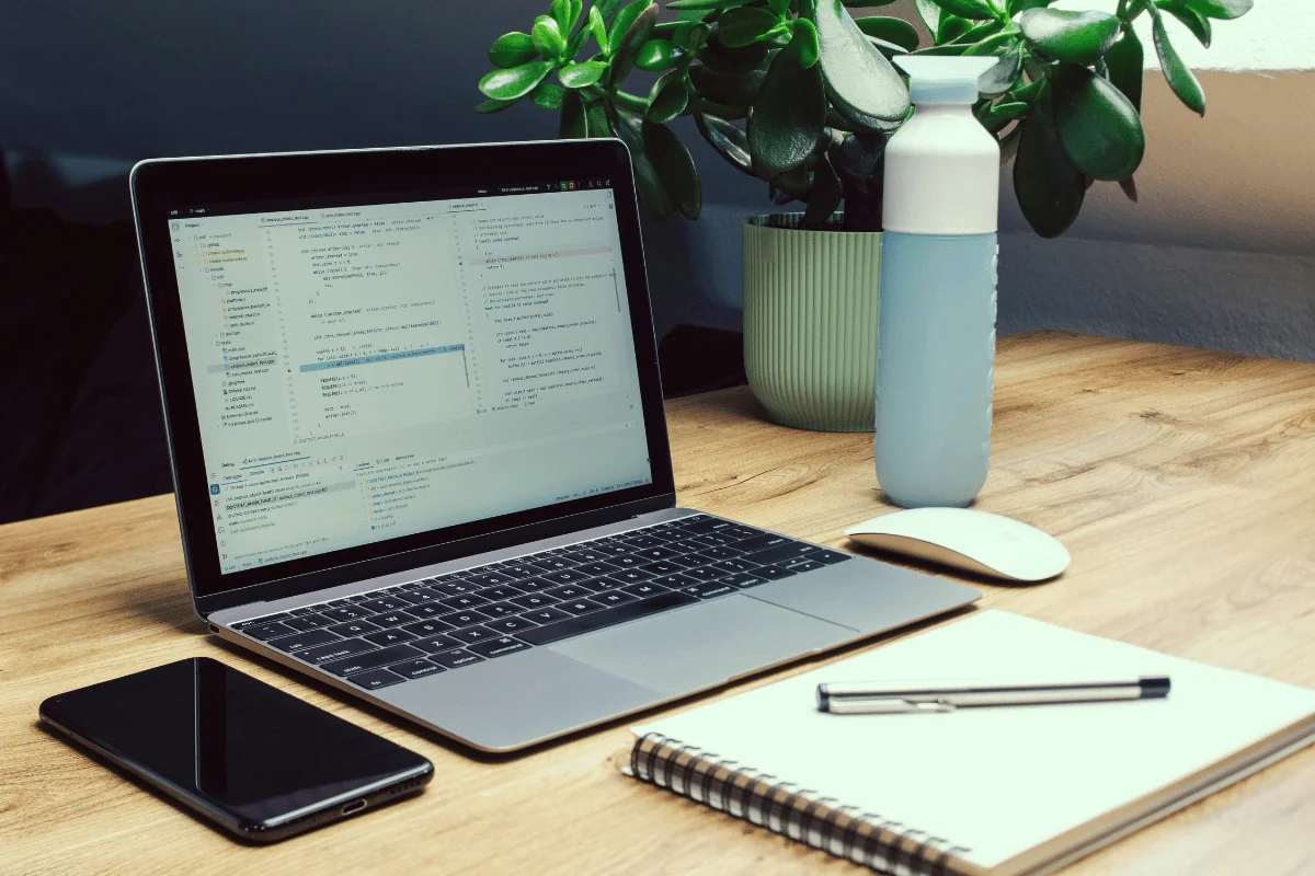 A laptop displaying code sits on a wooden desk alongside a smartphone, notebook, pen, mouse, and a green plant.