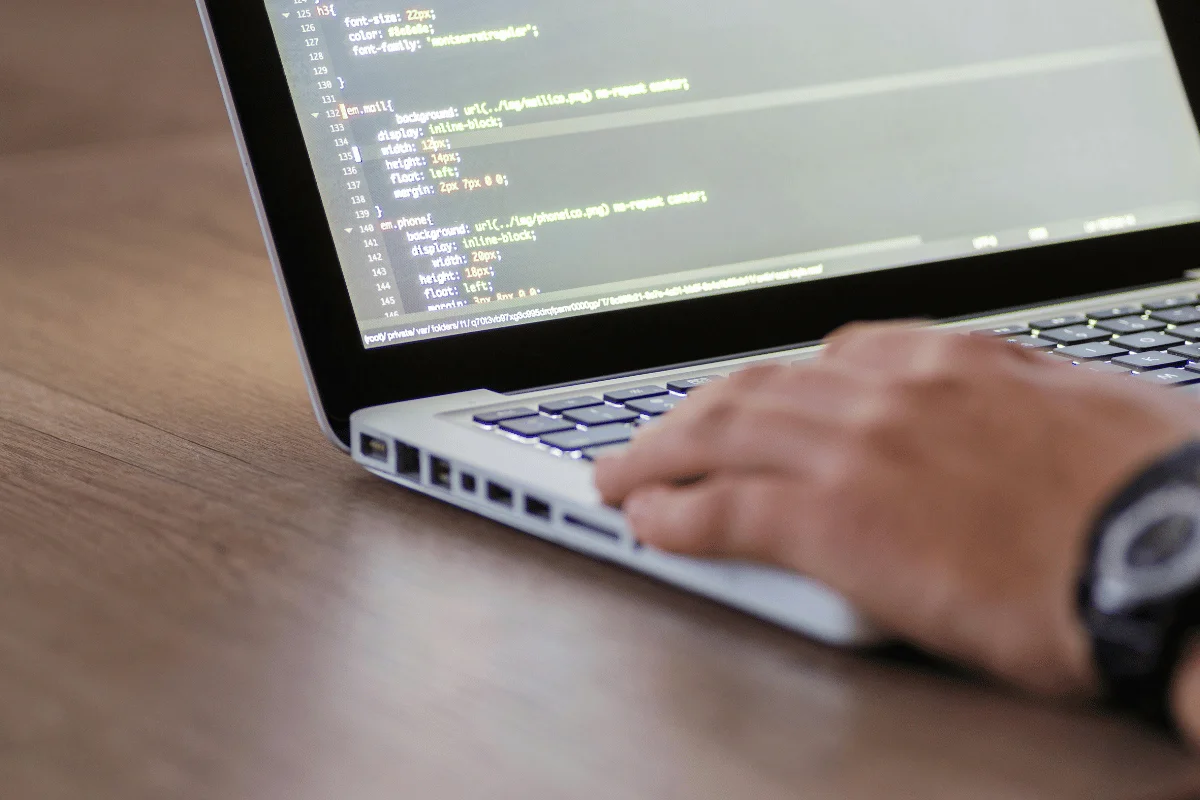 A close-up of a laptop keyboard with a hand typing code on the screen, showcasing lines of HTML and CSS programming.