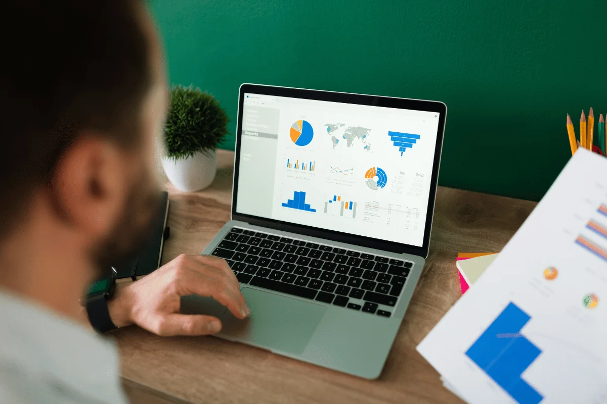 A person views colorful data visualizations on a laptop, surrounded by office supplies and a small plant on a wooden desk.
