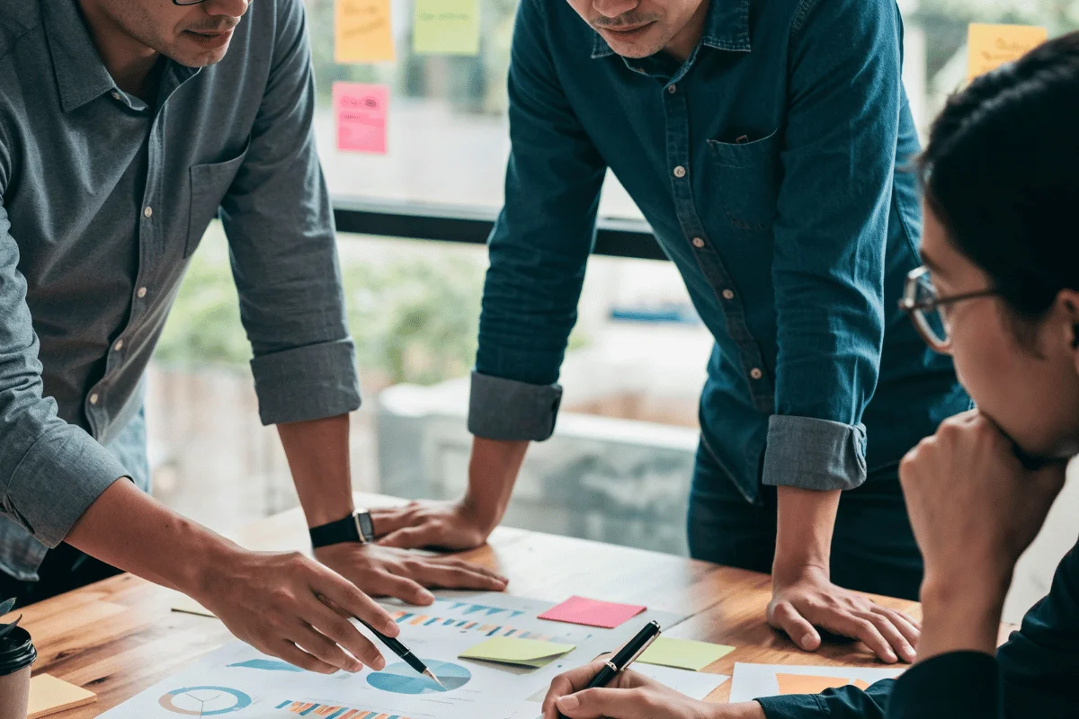 Three people stand around a table discussing a chart. The atmosphere is collaborative and focused, with colorful sticky notes visible in the background.