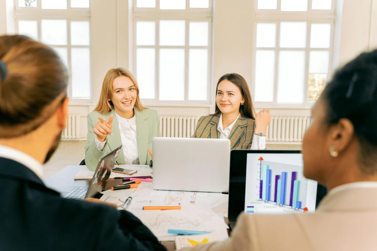 A group of women in business attire engaged in a meeting, discussing over laptops and charts on a table in a bright room.