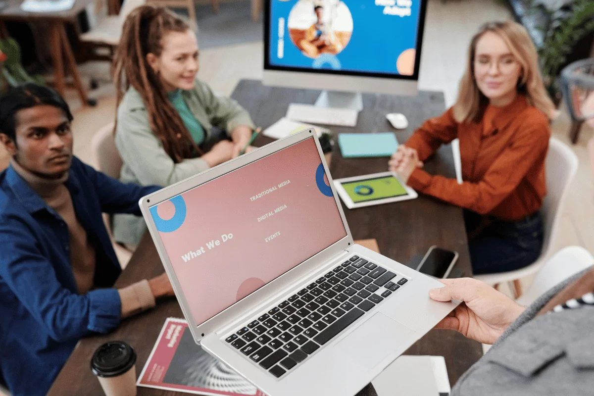 A laptop displays a presentation titled "What We Do," while a group of people engages in discussion at a table.