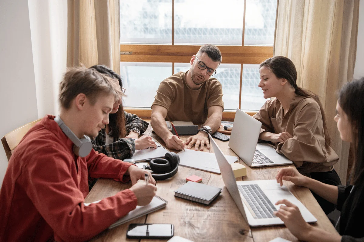 A group of people collaborate at a wooden table, filled with laptops, notebooks, and stationery, in a bright, casual workspace.