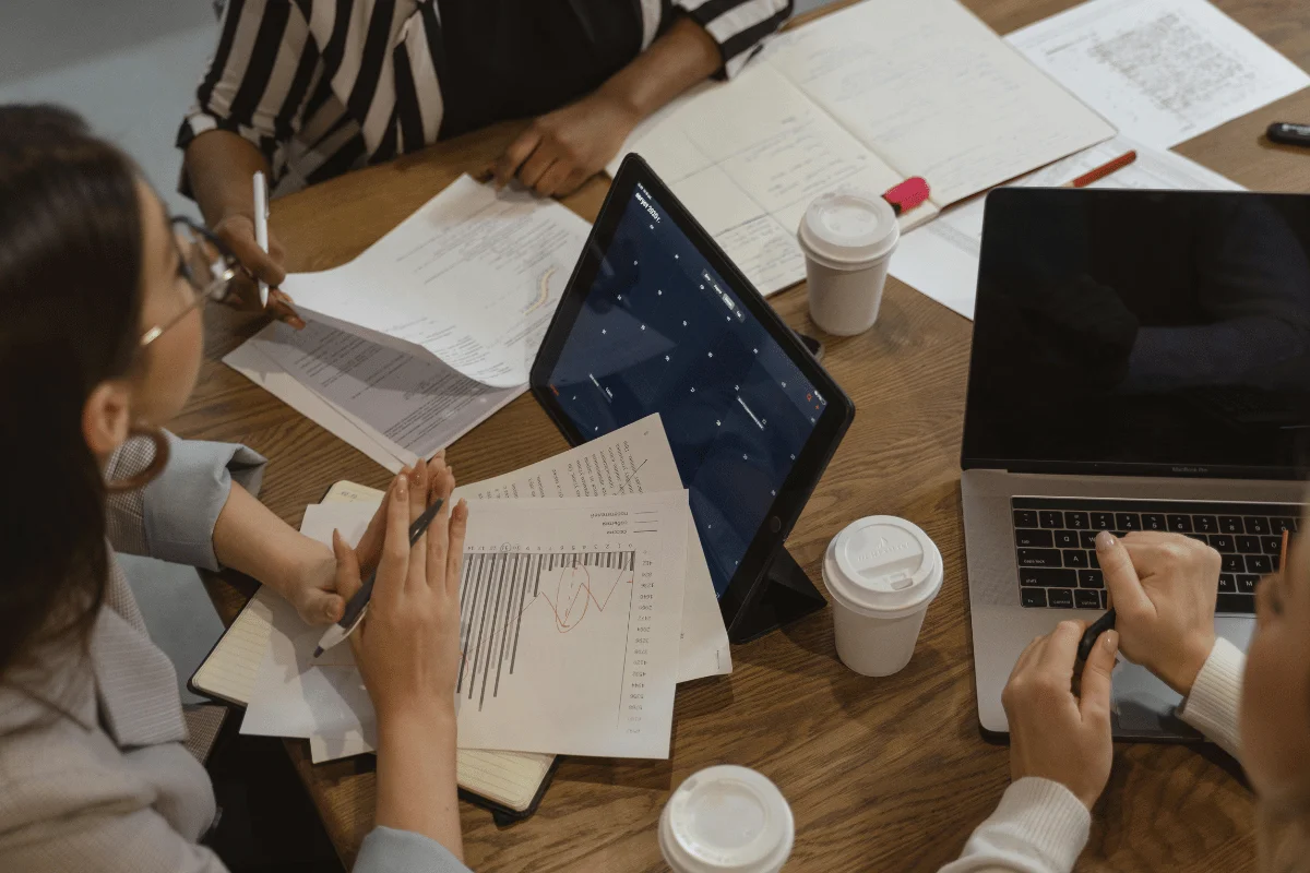 A table covered with papers, a laptop, a tablet displaying data, and coffee cups. People are engaged in discussion and note-taking.