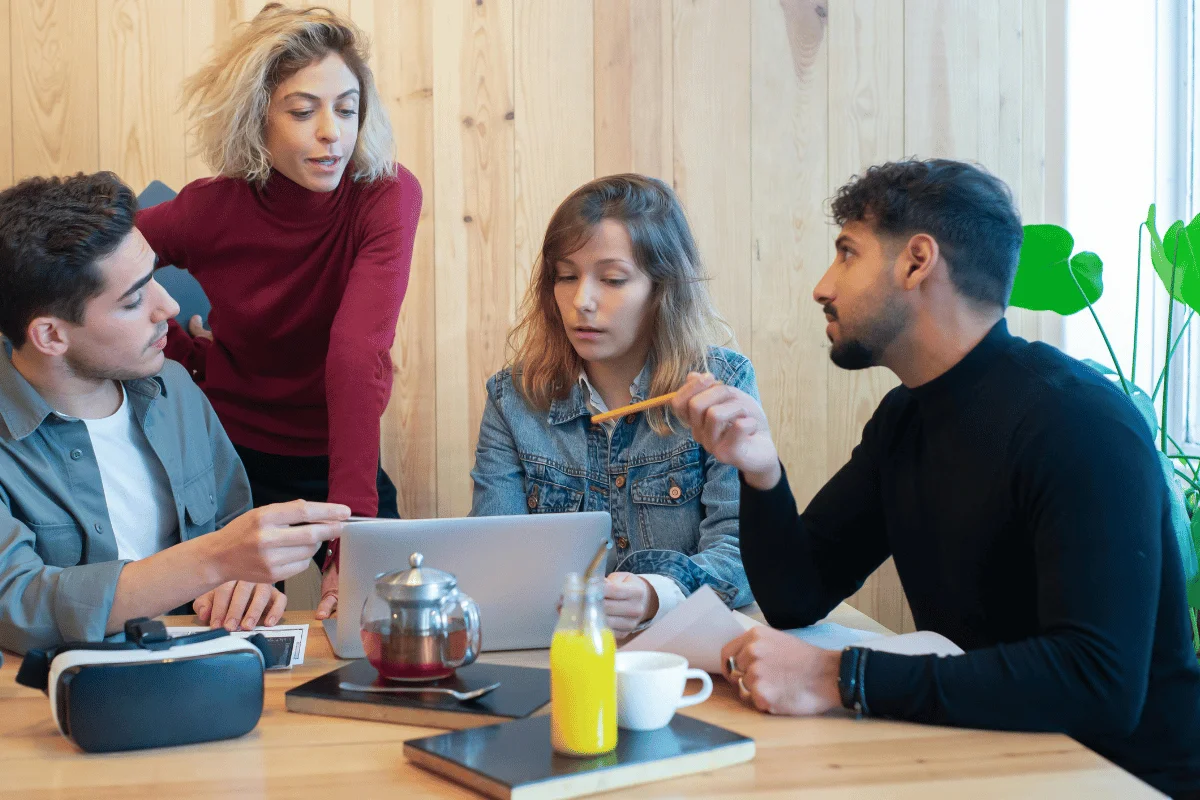 Four people engage in a discussion around a table with laptops and notebooks. The atmosphere is focused.