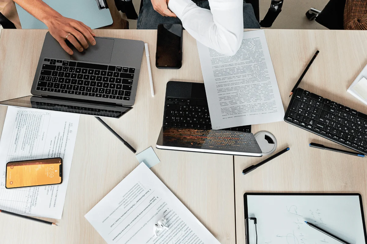 A top-down view of a work desk with two laptops, a smartphone, papers, pens, and a tablet, showcasing a busy workspace.